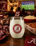 A white stemless wine glass candle with a red and white Alabama Crimson Tide logo, set on a wooden table in a lively football game厅 with fans, popcorn, and a pizza box in the background.