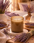 A lit candle in a speckled amber glass jar sits on a wooden table surrounded by lavender sprigs, bowls of dried lavender, and decorative vases in a warm, softly lit bedroom setting.