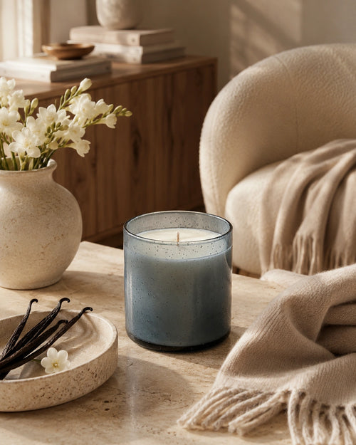 A blue-gray candle in a clear glass jar sits on a wooden table in a cozy, sunlit room with a cream chair, throw blanket, flowers in a vase, and vanilla beans.