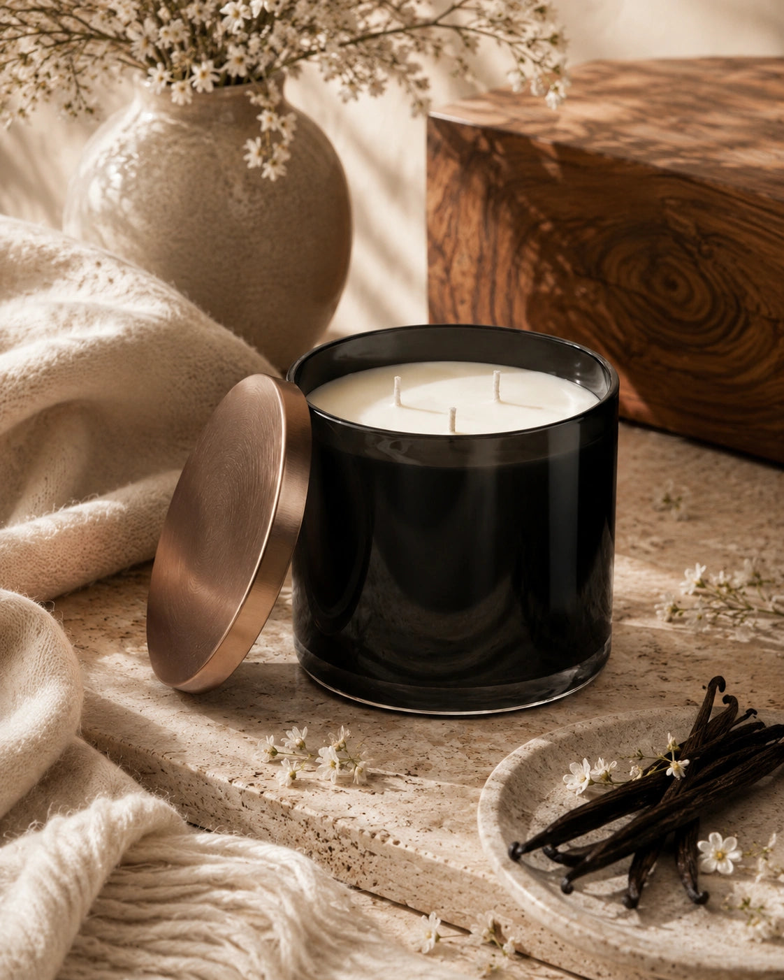 A black glass candle jar with a metallic rose-gold lid leaning beside it, set on a stone surface with cream knit fabric, dried flowers, vanilla pods, and a wooden block in the background.