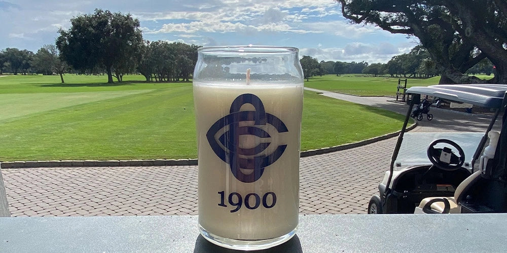 Candle in a glass container with a logo and '1900' text, set against a golf course background.