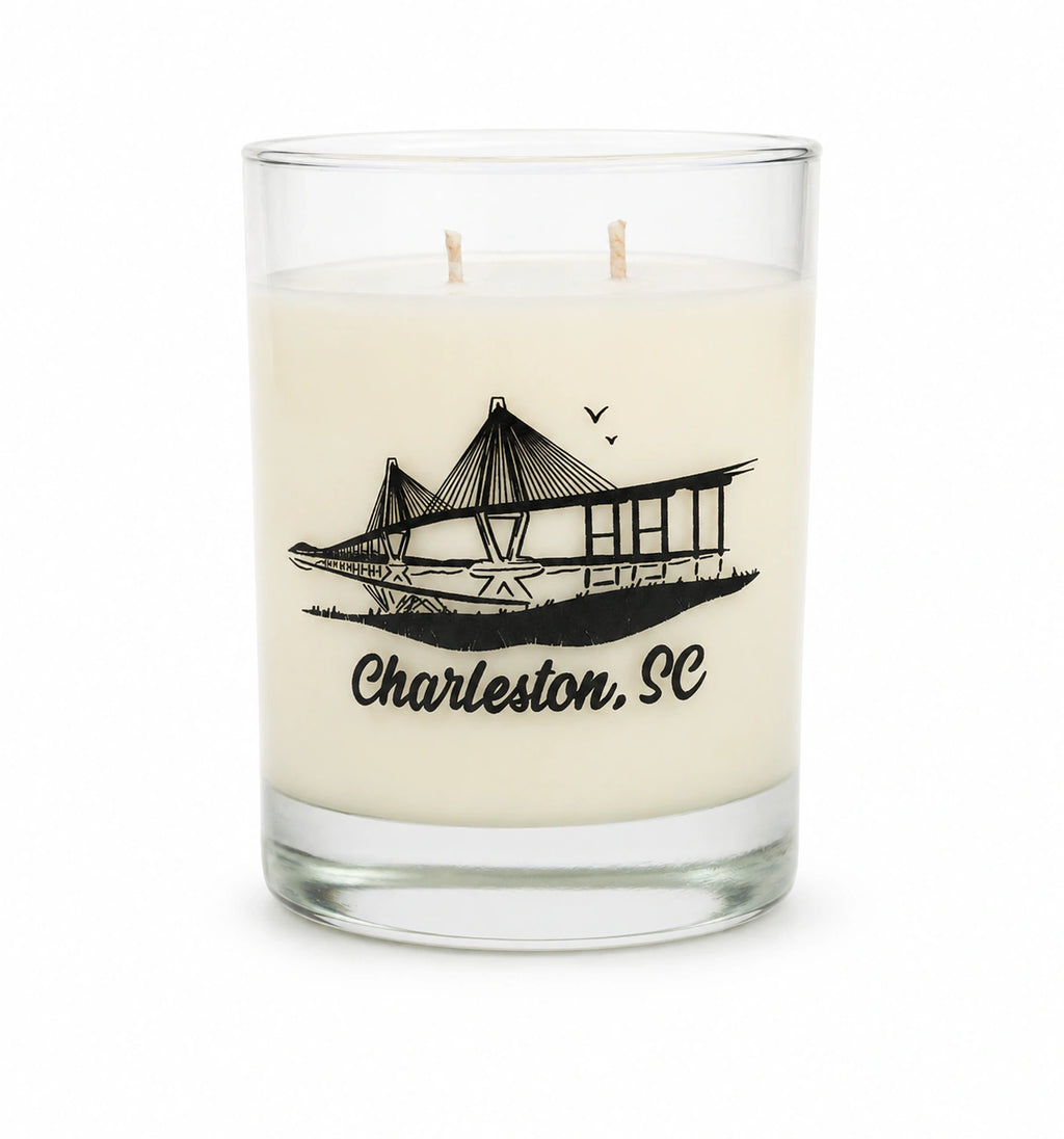 An off-white scented candle in a clear glass jar with two wicks, featuring a black graphic of the Charleston, SC bridge and the text 'Charleston, SC' printed on the front.