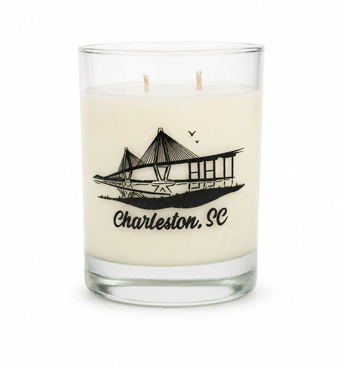 An off-white scented candle in a clear glass jar with two wicks, featuring a black graphic of the Charleston, SC bridge and the text 'Charleston, SC' printed on the front.