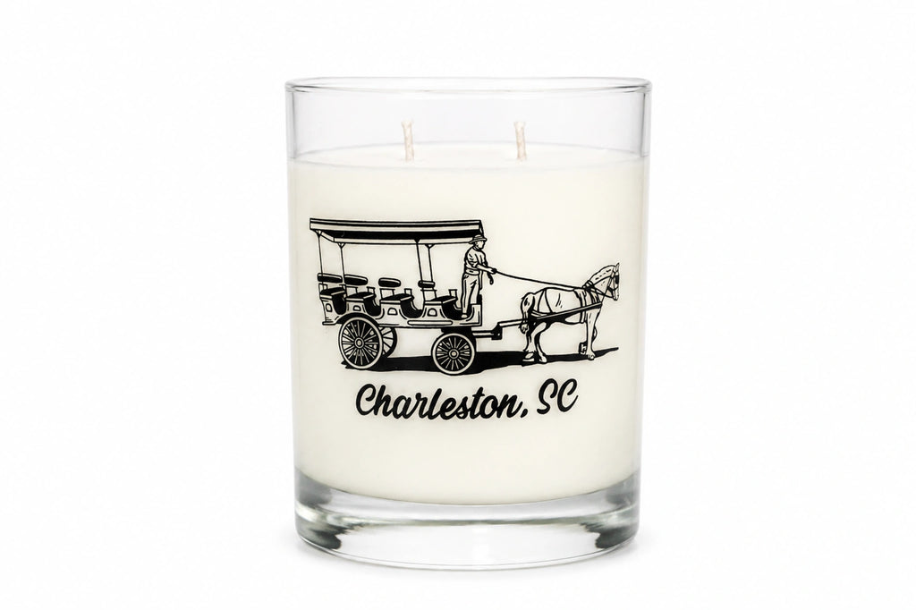A white wax candle in a clear glass jar with two wicks, featuring a black graphic of a horse-drawn carriage and the text 'Charleston, SC' on the front, set against a plain white background.