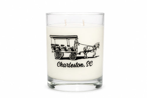 A white wax candle in a clear glass jar with two wicks, featuring a black graphic of a horse-drawn carriage and the text 'Charleston, SC' on the front, set against a plain white background.