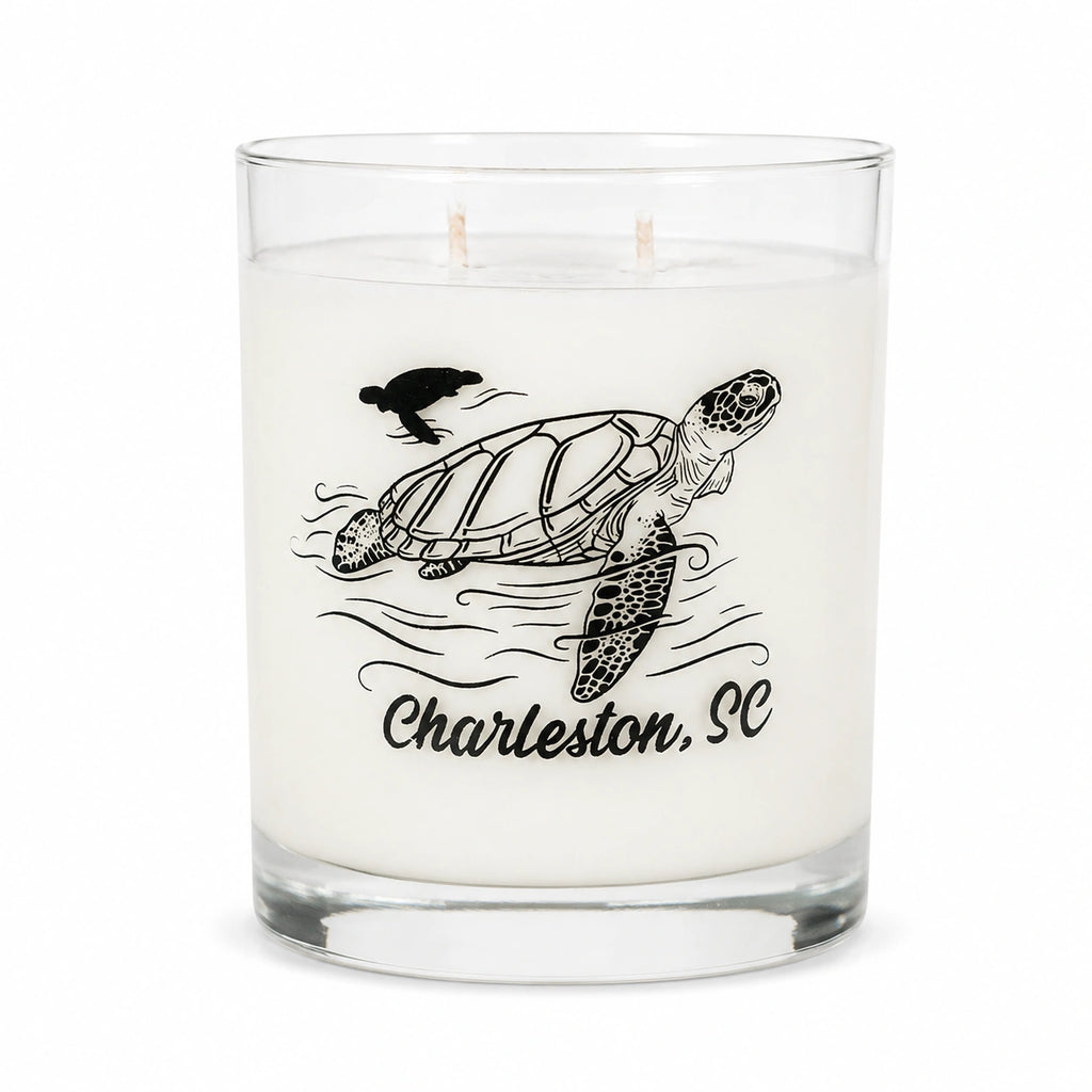 A white scented candle in a clear glass jar with two wicks, featuring a black line art illustration of a sea turtle, waves, a bird, and the text 'Charleston, SC' on the front.