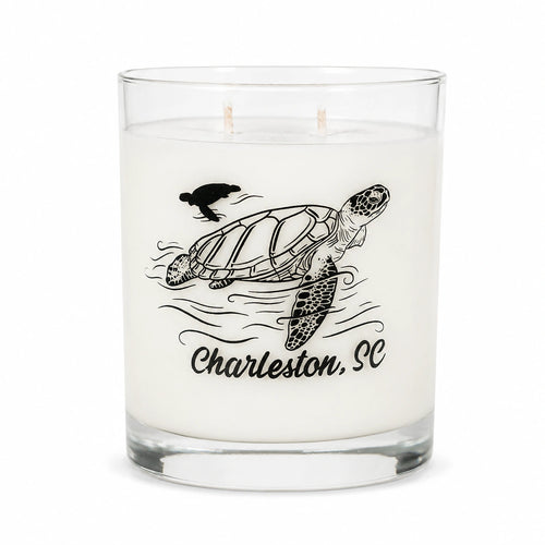A white scented candle in a clear glass jar with two wicks, featuring a black line art illustration of a sea turtle, waves, a bird, and the text 'Charleston, SC' on the front.