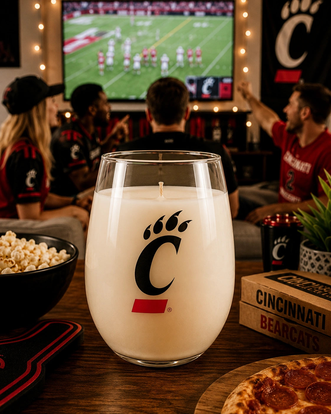A large candle in a clear stemless glass with a Cincinnati Bearcats logo, set on a wooden table in a bar with people watching a football game in the background.