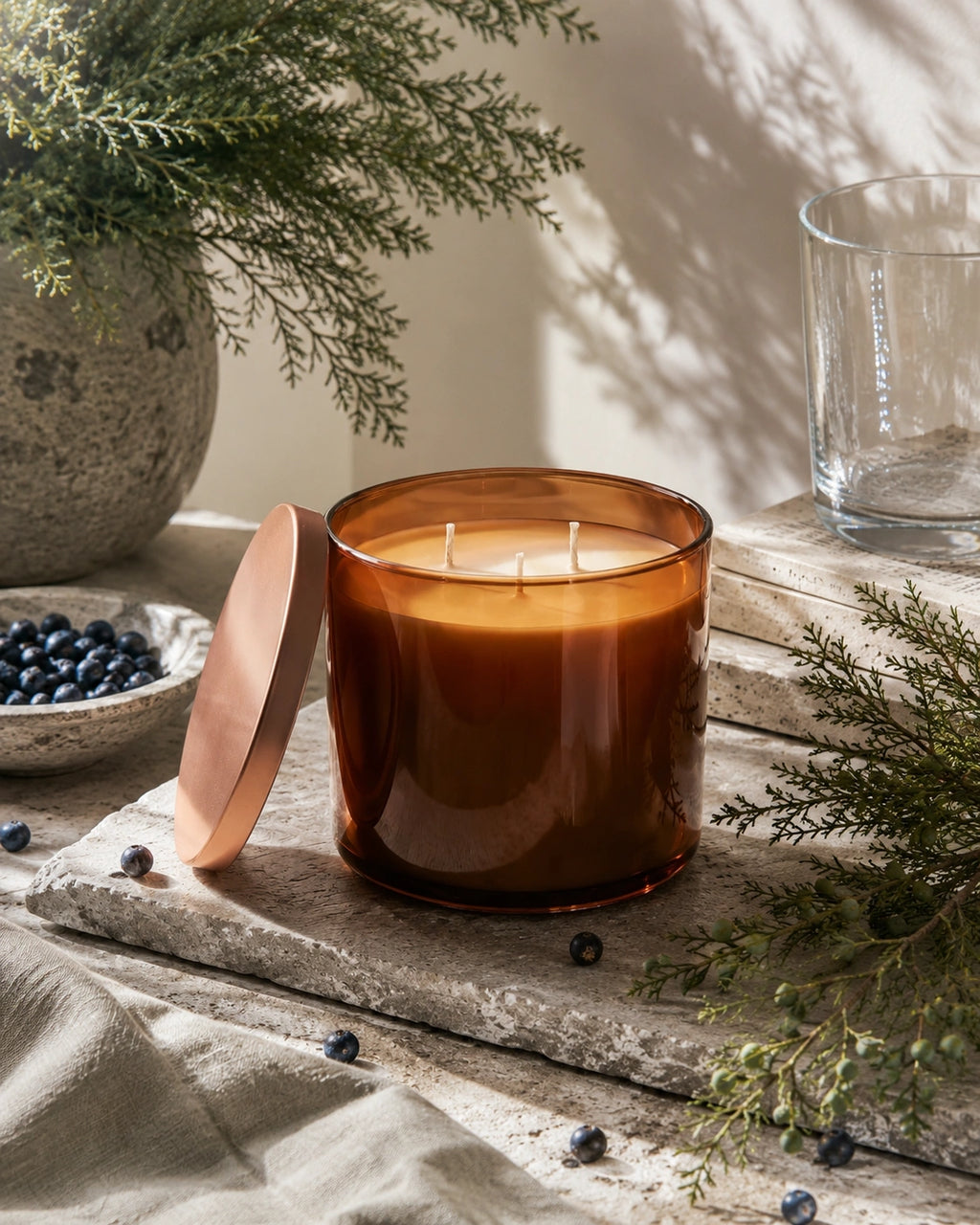 An amber glass candle jar with two wicks sits on a rustic stone surface, with its matching lid leaning beside it. The scene includes evergreen branches, blueberries, a clear glass tumbler, and soft natural shadows, creating a styled product presentation.