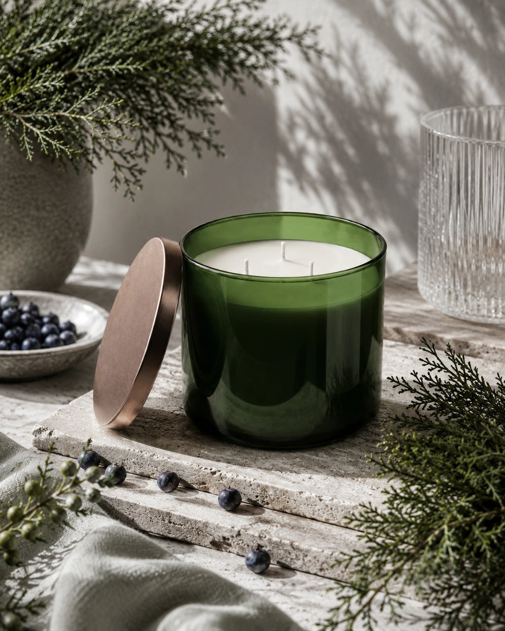A green glass candle jar with its metallic rose-gold lid leaning beside it, styled on a rustic stone surface with evergreen branches, blueberries, and a ribbed glass tumbler in soft natural light.