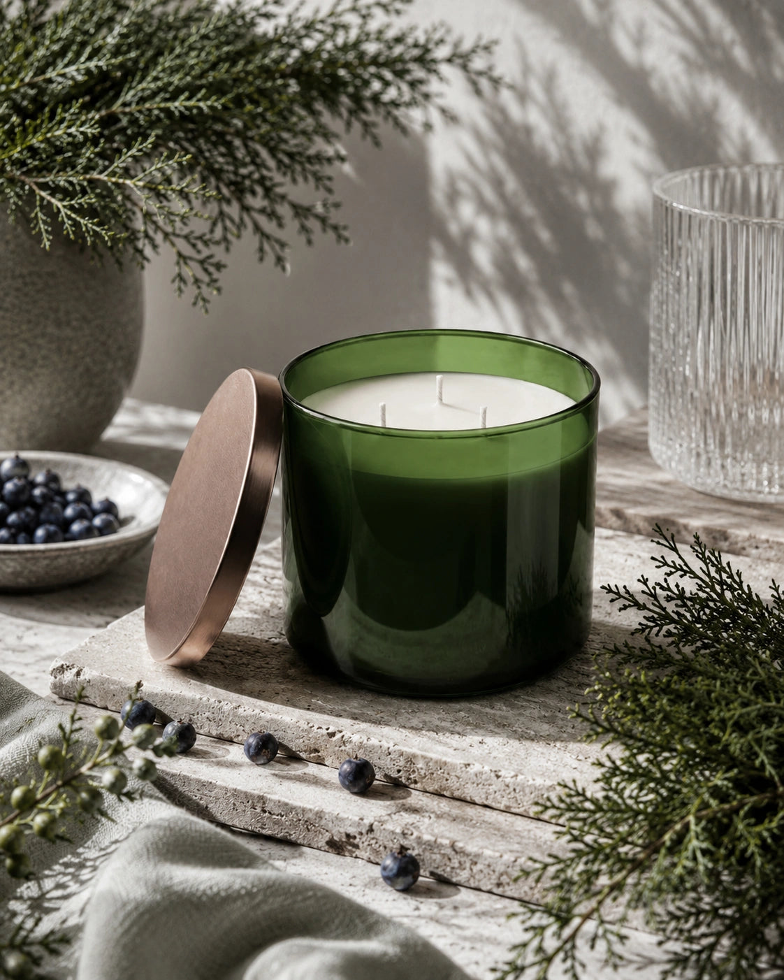 A green glass candle jar with its metallic rose-gold lid leaning beside it, styled on a rustic stone surface with evergreen branches, blueberries, and a ribbed glass tumbler in soft natural light.