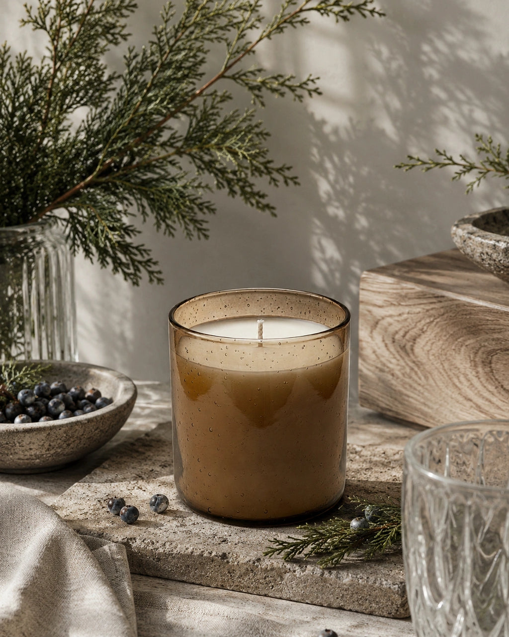 A lit candle in a speckled amber glass jar sits on a rustic stone surface with pine branches, blueberries, and neutral-toned props in a softly lit indoor setting.