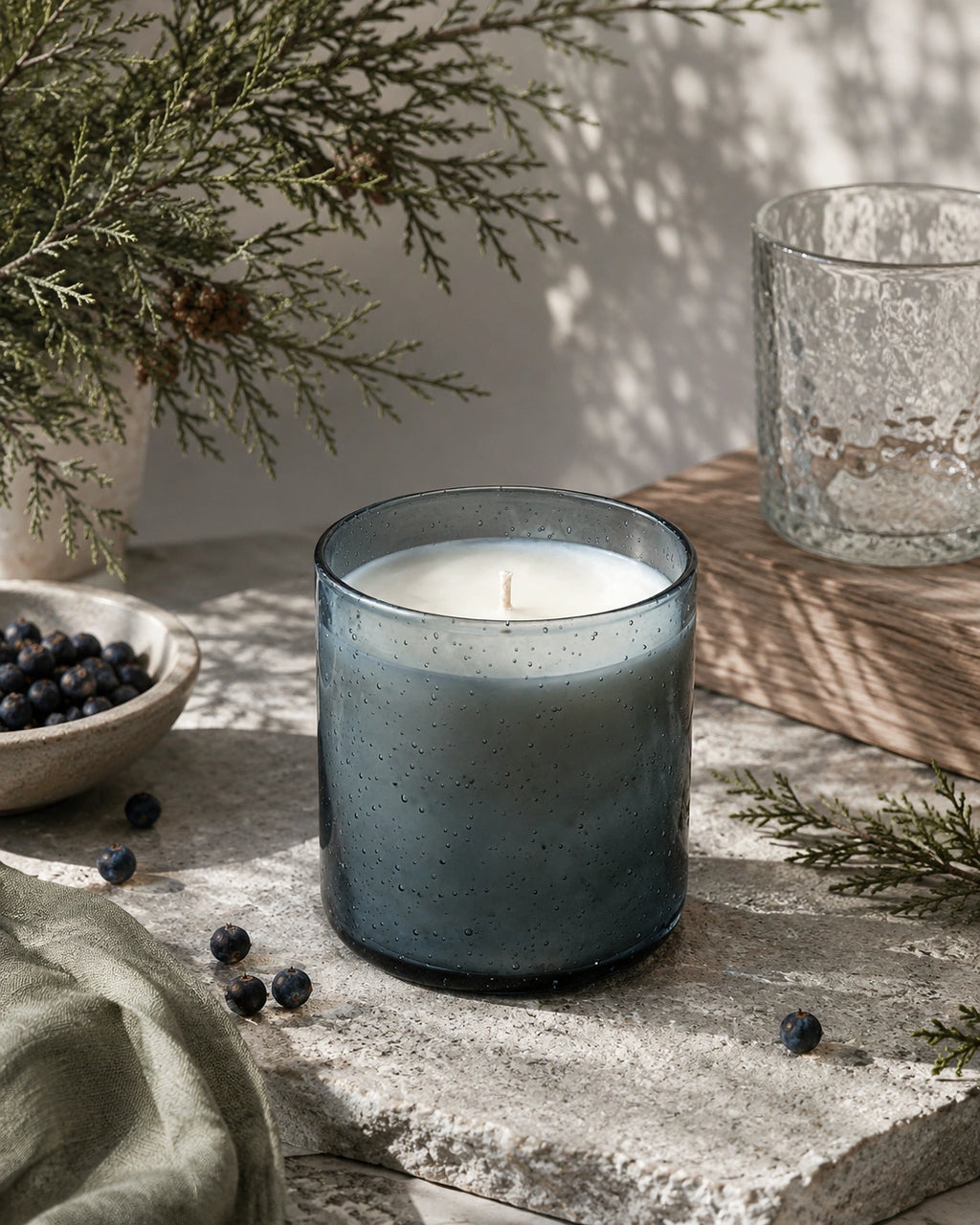A lit candle in a speckled smoky blue-gray glass jar sits on a stone surface with blueberries, evergreen branches, and a textured glass vessel in a softly lit indoor setting.