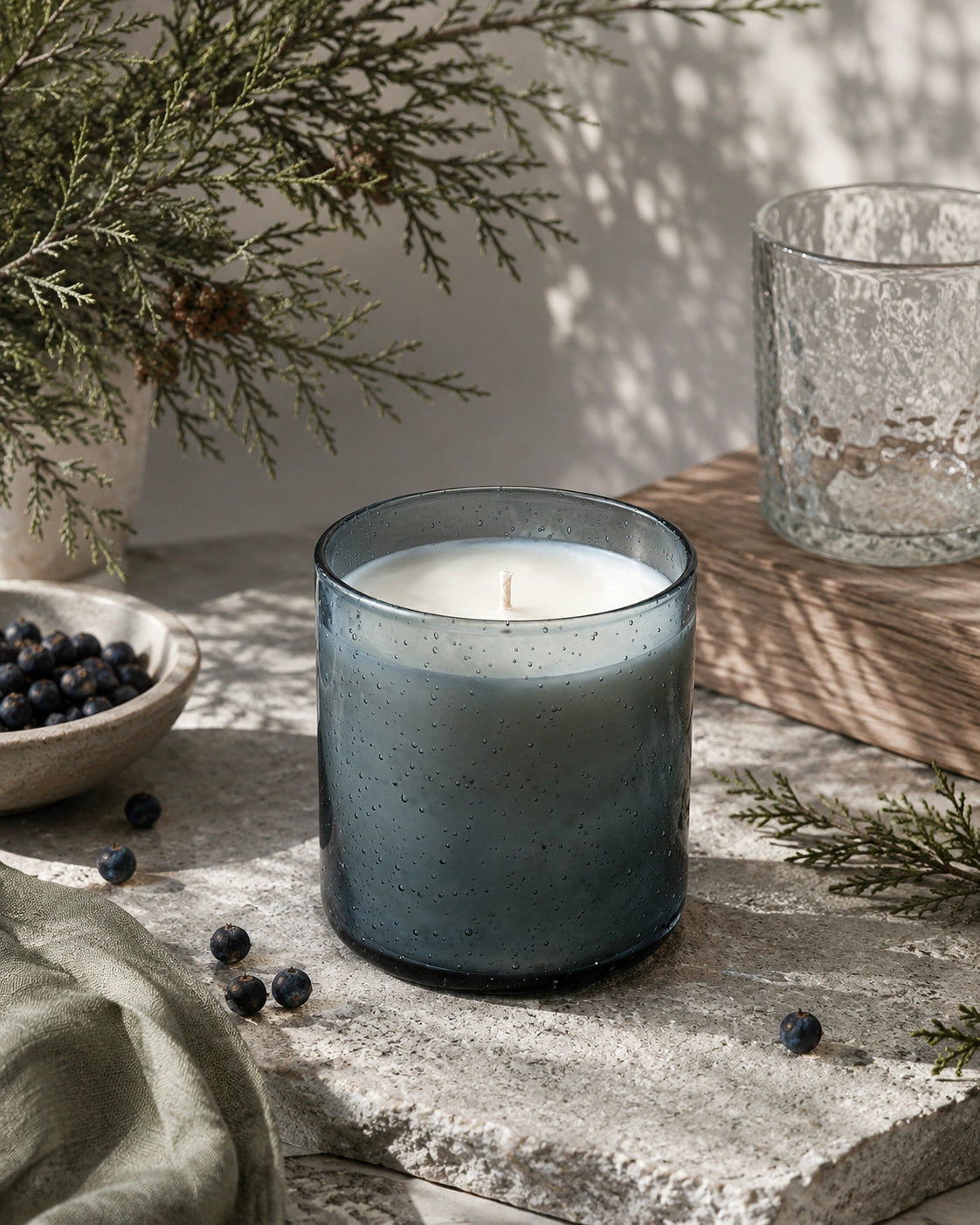 A lit candle in a speckled smoky blue-gray glass jar sits on a stone surface with blueberries, evergreen branches, and a textured glass vessel in a softly lit indoor setting.
