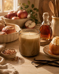 A lit candle in a smoky amber glass jar sits on a wooden table surrounded by autumn food props, including apples, baked goods, vanilla beans, a bottle of amber liquid, and a ceramic utensil holder.