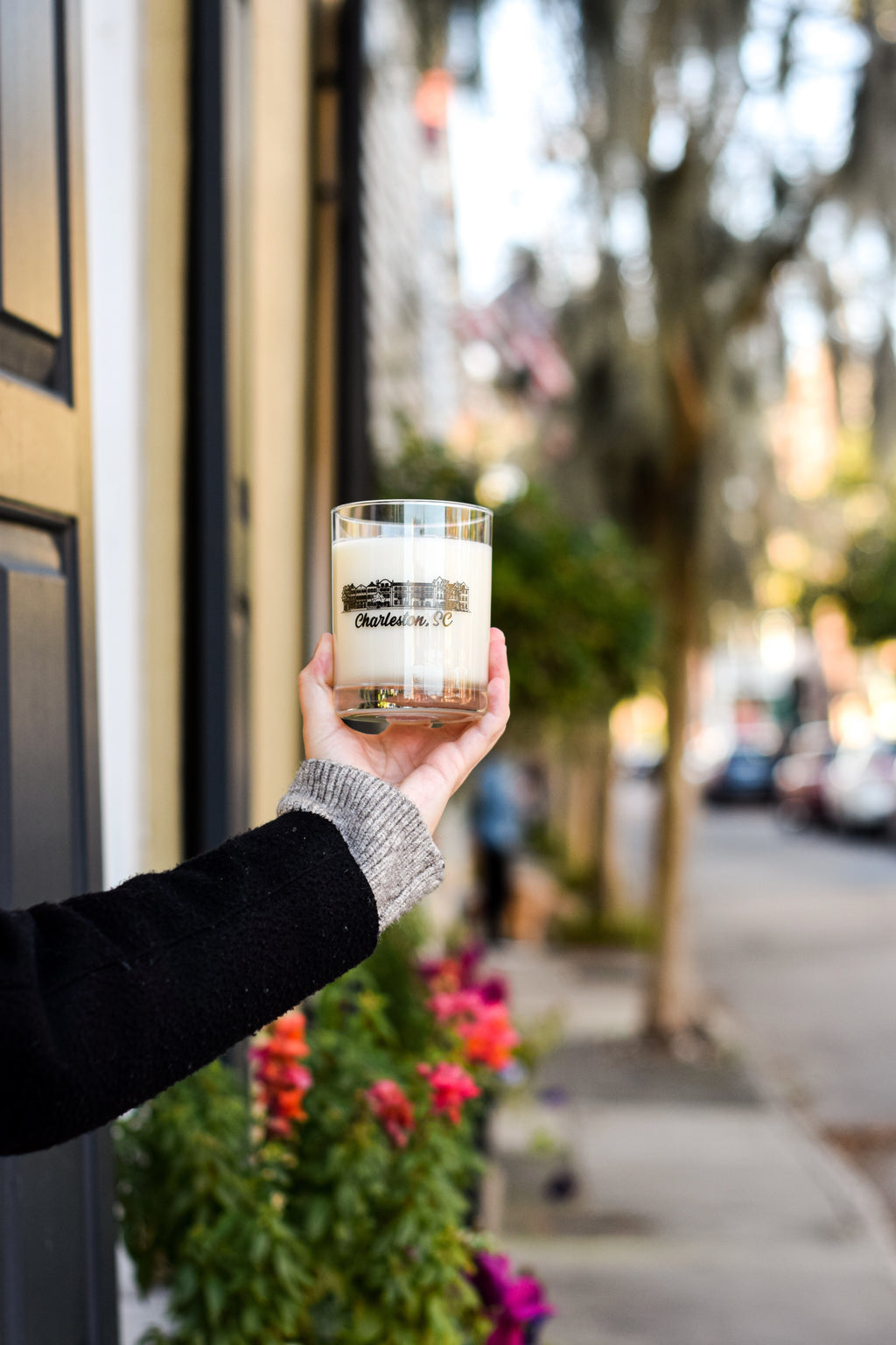 A hand holding a clear glass jar candle with a cream-colored wax and a black label that reads 'Belle Isle'. The background is a softly blurred outdoor street scene with trees, parked cars, and flowers.