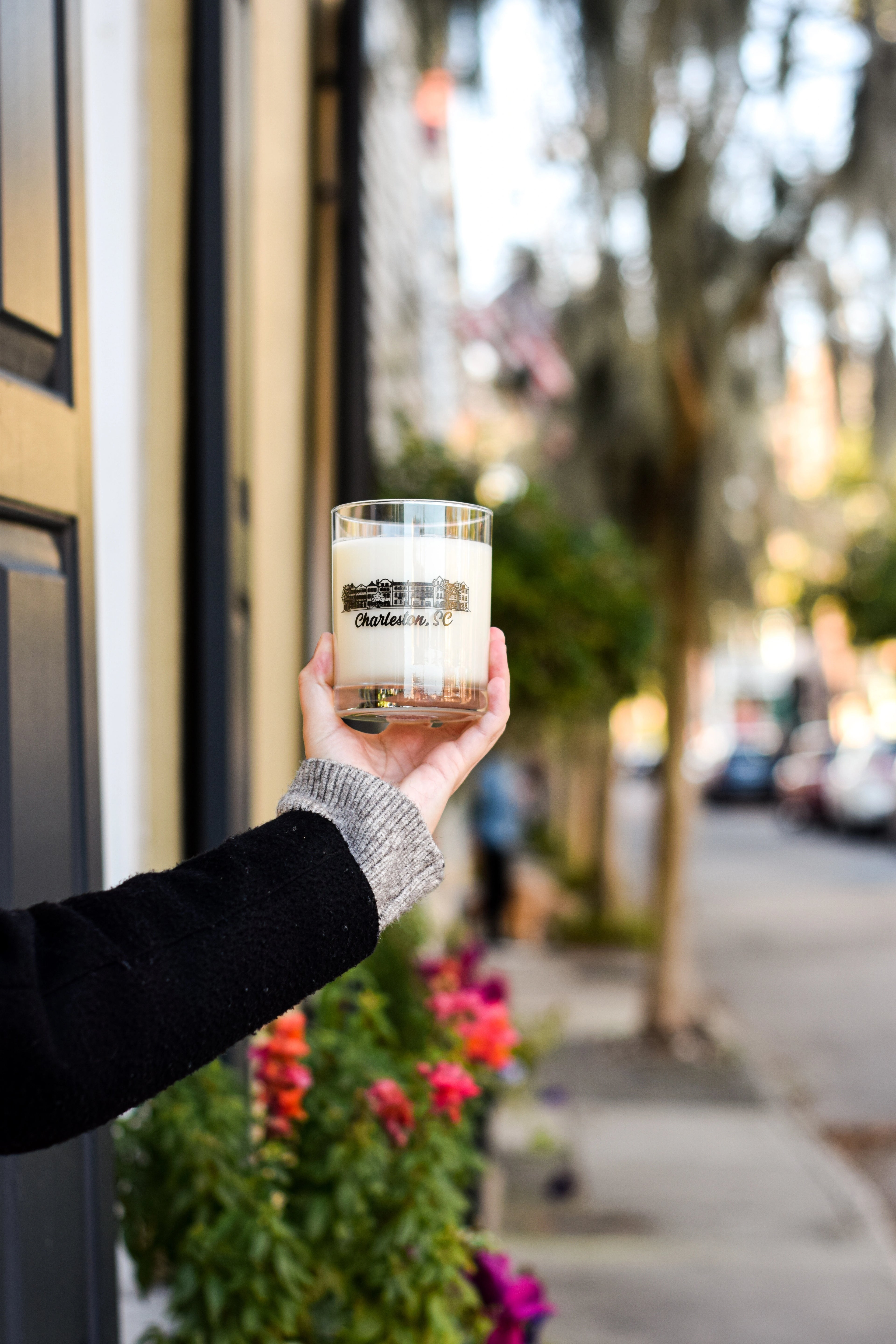 A hand holding a clear glass jar candle with a cream-colored wax and a black label that reads 'Belle Isle'. The background is a softly blurred outdoor street scene with trees, parked cars, and flowers.
