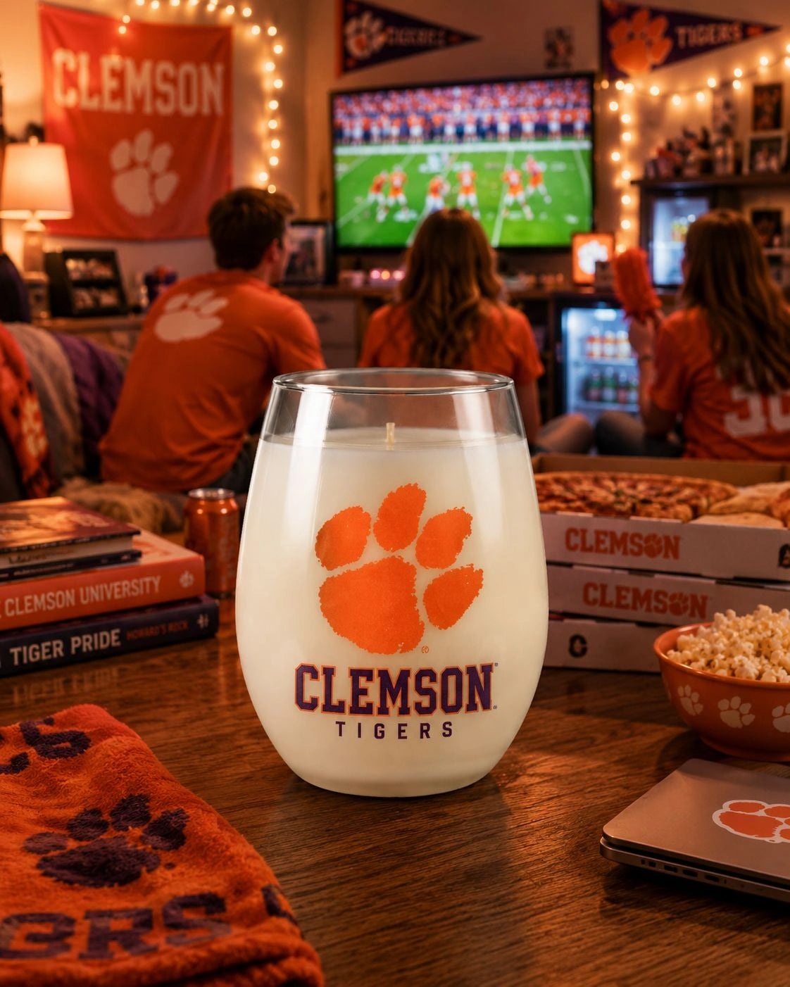 A Clemson Tigers stemless wine glass with an orange paw print logo and 'Clemson Tigers' text, filled with white wine and set on a wooden table in a lively bar with fans watching a football game in the background.