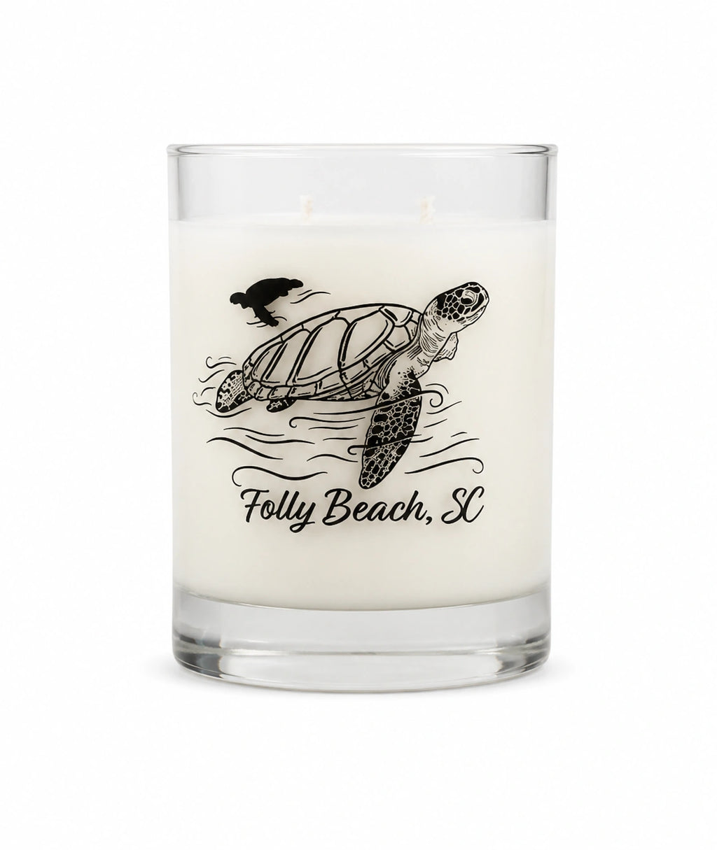 A white scented candle in a clear glass jar with a black illustration of a sea turtle, bird, and waves, with the text 'Folly Beach, SC' printed below.