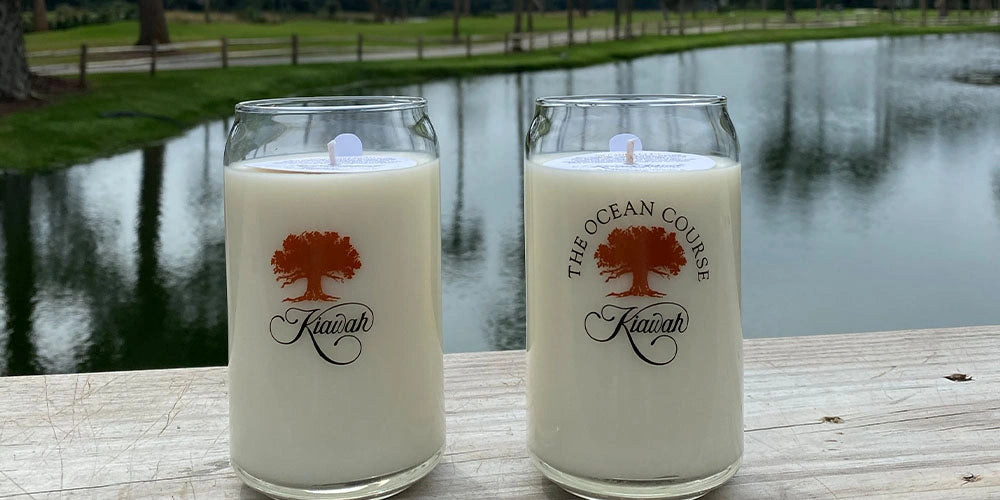 Two candles with 'The Ocean Course Kawah' branding on a wooden surface near a body of water.
