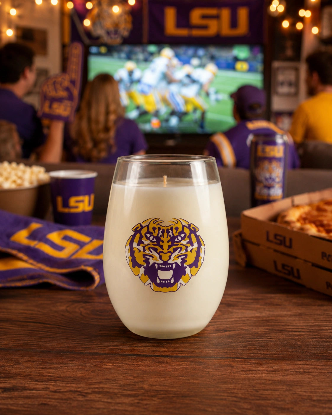 A wine-stemless glass candle with a white wax fill and a purple and gold LSU tiger logo on the front, set on a wooden table in a lively bar or lounge with people watching a football game in the background.