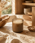 A lit candle in a frosted glass jar sits on a stone slab in a warm, rustic interior setting with wooden shelves, dried grass, and natural light.