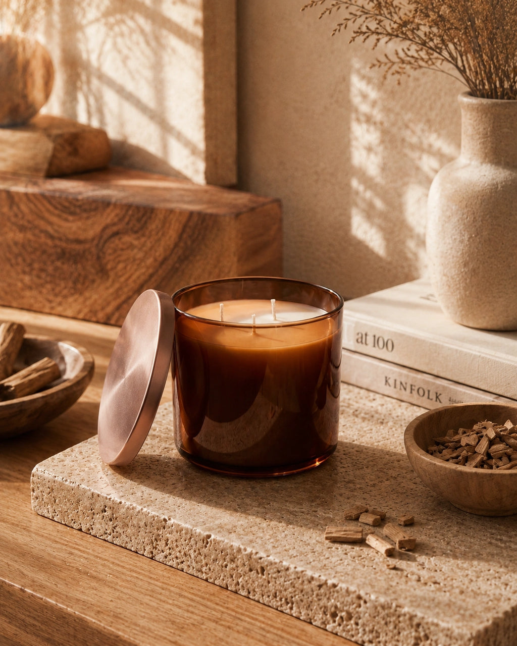 An amber glass candle jar with its matching lid leaning beside it on a stone slab, styled in a warm rustic room with sunlight, books, a vase, and small bowls.