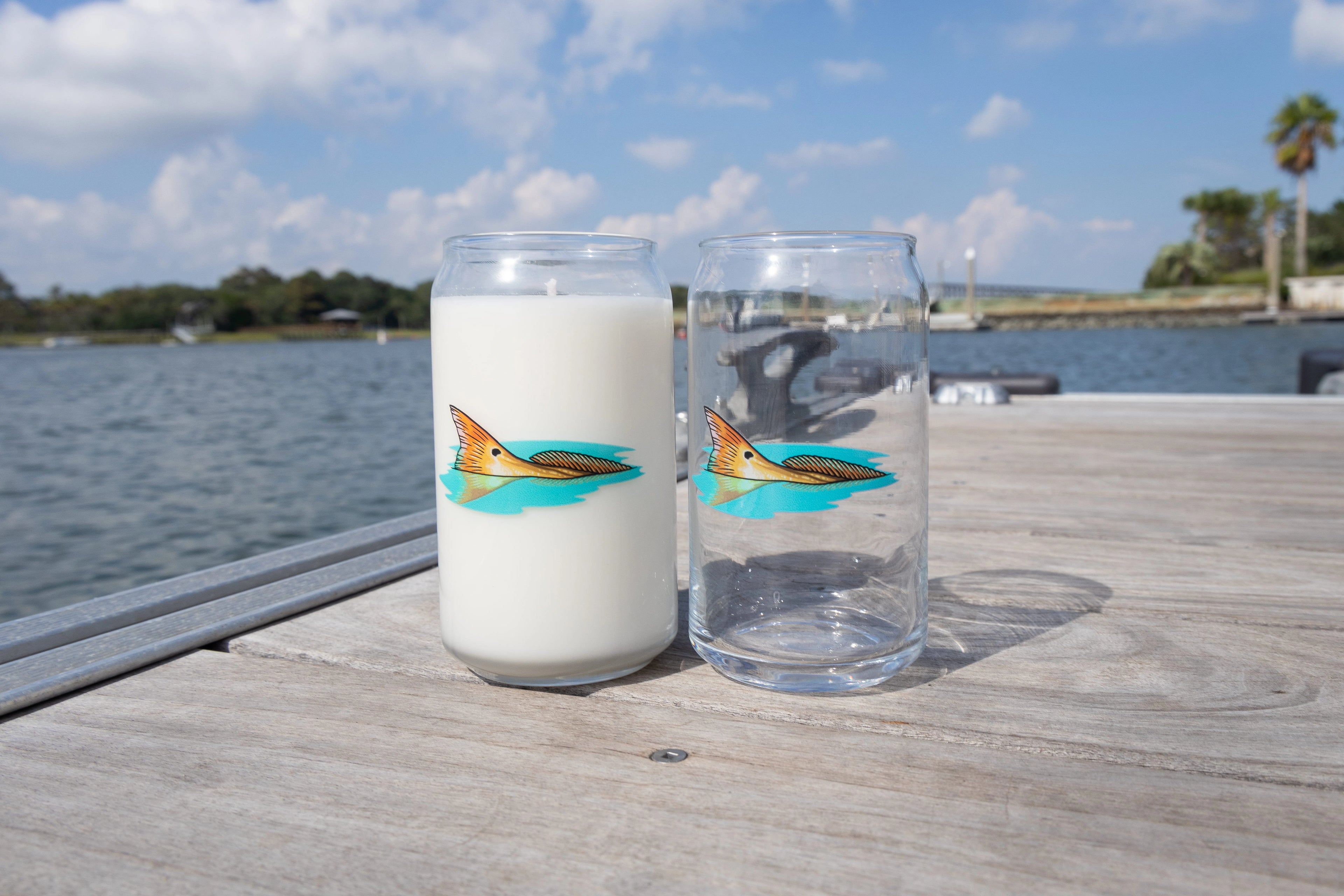 Two glasses with fish illustrations on a wooden dock by a body of water.