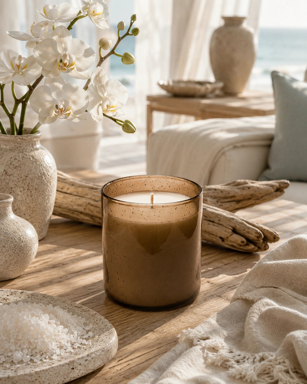 A brown amber glass jar candle sits on a wooden table in a beautifully styled, sunlit room with white orchids, ceramic vases, driftwood, and a cozy throw blanket.