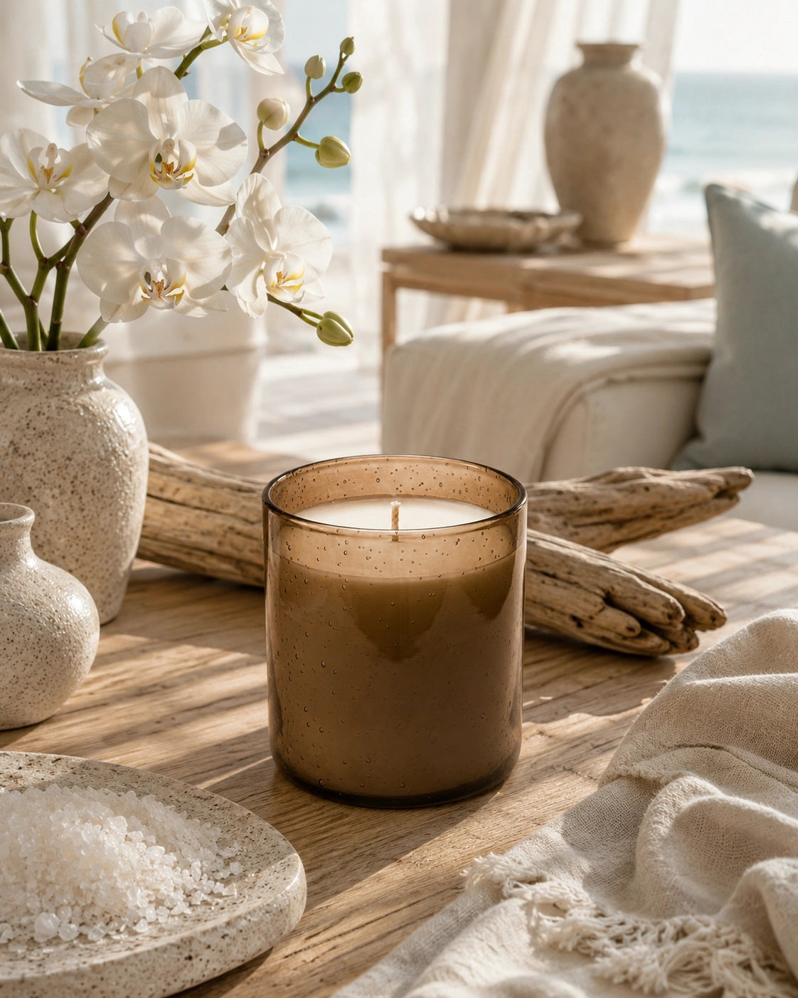 A brown amber glass jar candle sits on a wooden table in a beautifully styled, sunlit room with white orchids, ceramic vases, driftwood, and a cozy throw blanket.