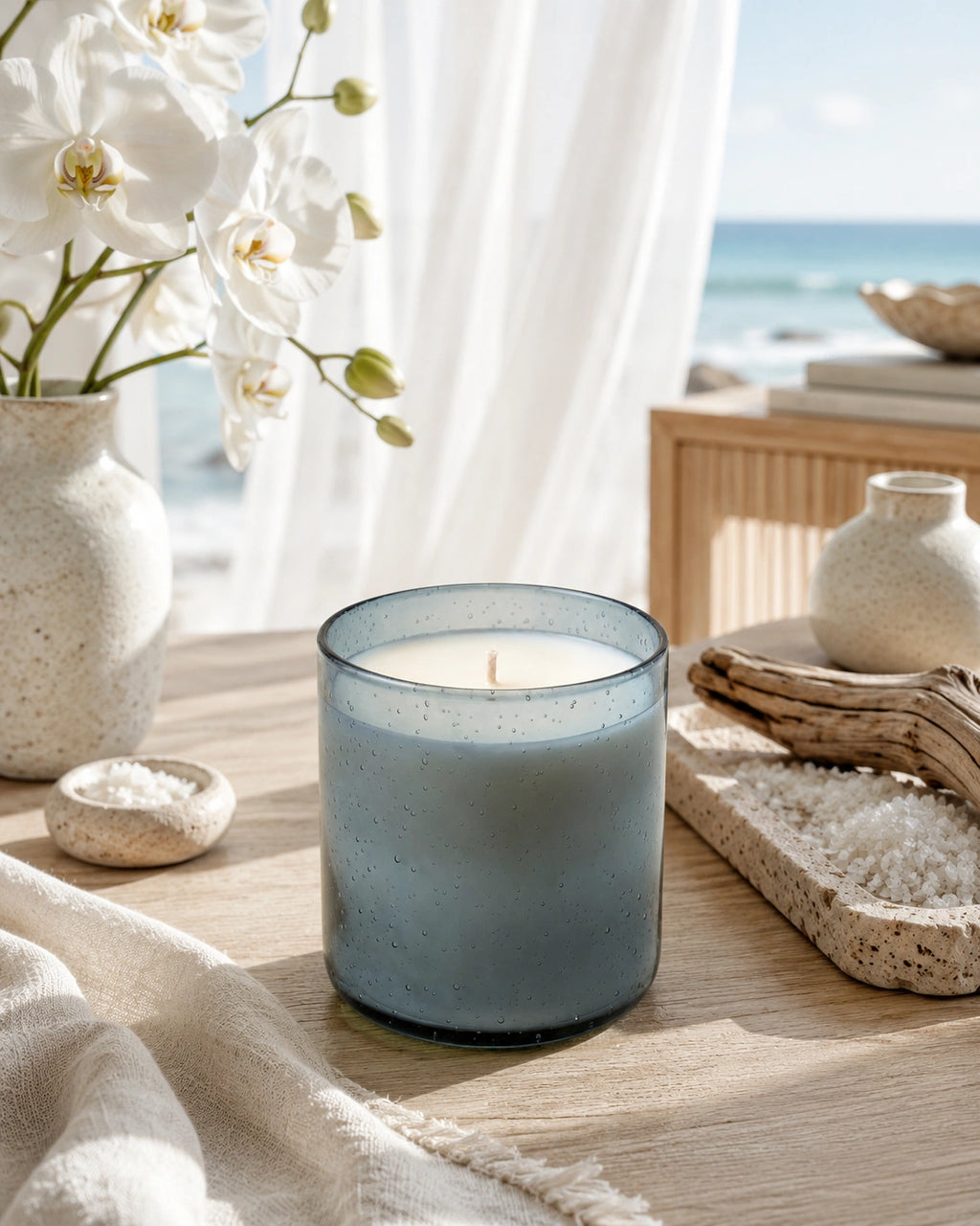 A lit candle in a speckled translucent blue-gray glass jar sits on a wooden table in a styled beach-side setting with white flowers, a vase, and ocean scenery in the background.