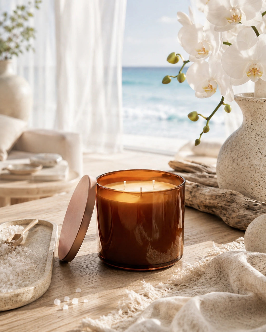 An amber glass jar candle with its lid leaning beside it on a wooden table, styled with white flowers, a textured vase, and a beach scene in the background.