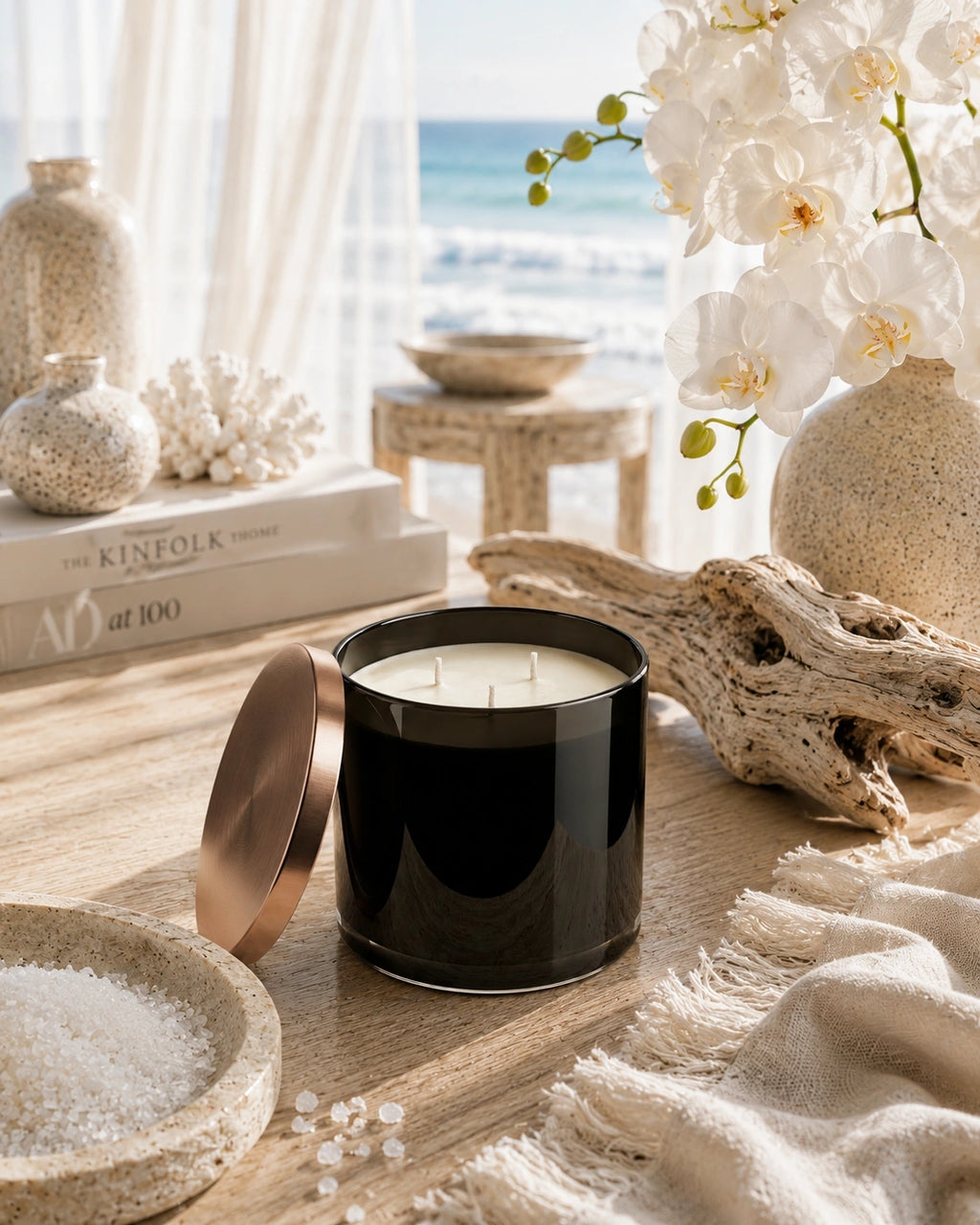 A black glass jar candle with a rose gold lid resting beside it on a wooden table, styled with white flowers, driftwood, a bowl of sea salt, and a beach view in the background.