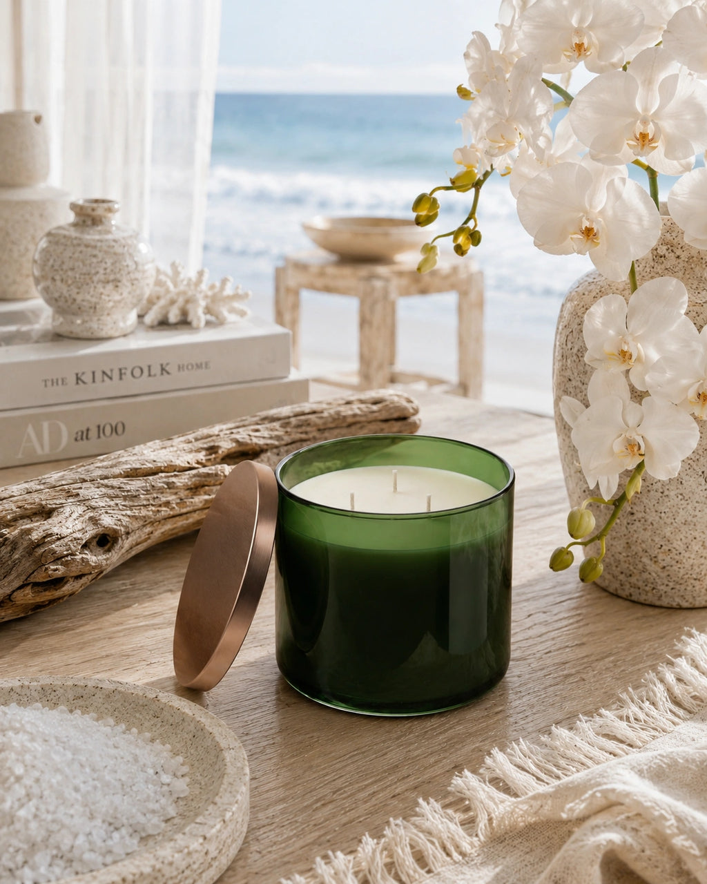 A dark green glass candle jar with its metallic rose-gold lid leaning beside it on a wooden table, styled with white orchids in a vase, driftwood, a plate of sea salt, and a beach scene in the background.