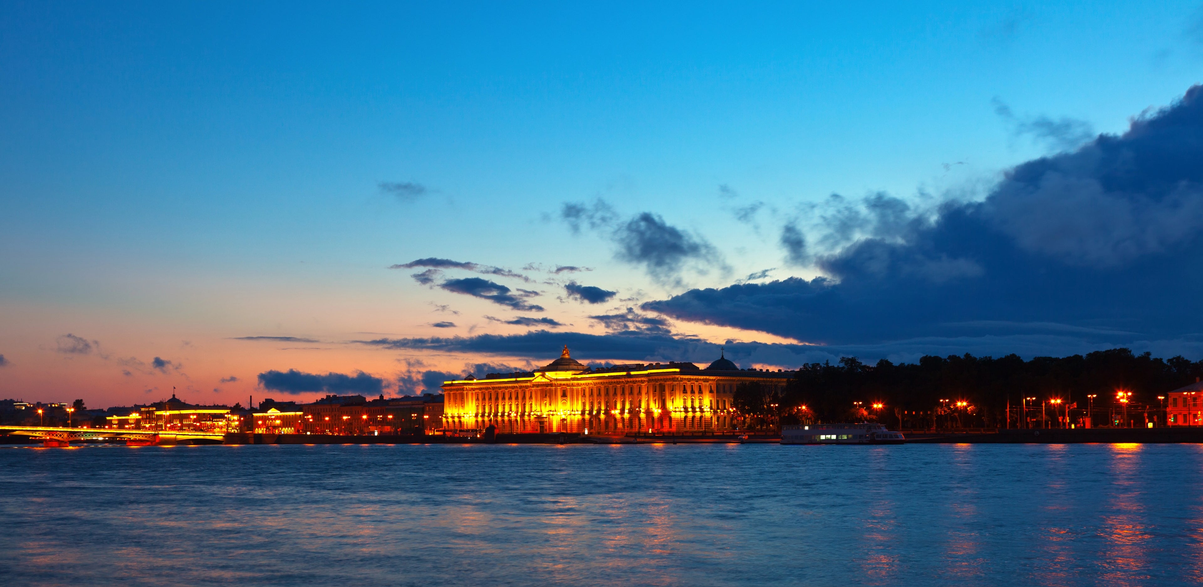 A scenic twilight photograph of a large illuminated building along the water, with reflections on the surface below and a blue sky with dark clouds above.
