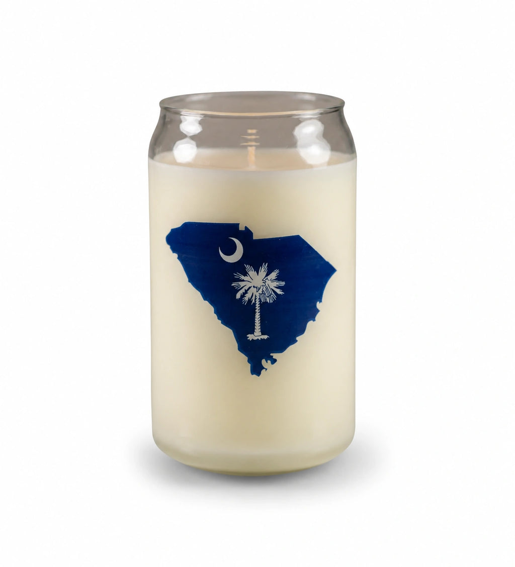 A jar candle with cream-colored wax inside a clear glass container, featuring a blue silhouette of South Carolina with a white palmetto leaf and crescent moon design on the front.