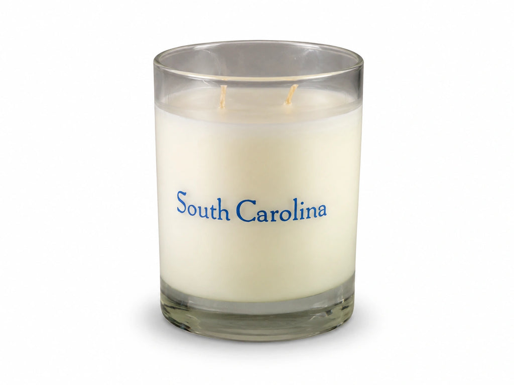 A single-wick scented candle in a clear glass jar with cream-colored wax and the words 'South Carolina' printed in blue on the front, set against a plain white background.