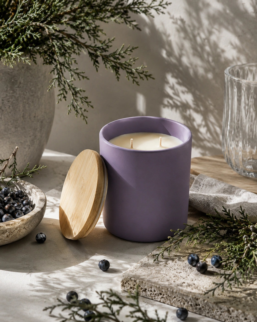 A matte lavender purple candle jar with a light natural wood lid leaning beside it on a styled tabletop. The scene includes evergreen branches, blueberries, a textured stone planter, a clear glass tumbler, and soft shadow lighting against a neutral wall.