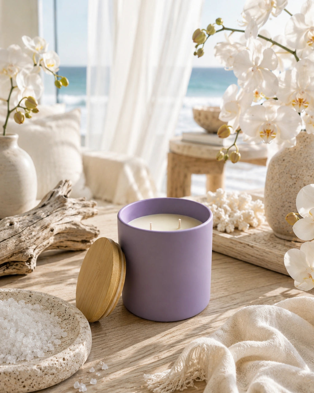 A lavender purple candle jar with a light wood lid resting beside it on a wooden table, styled with white orchids, driftwood, a bowl of salt, and a cozy beach-view background.