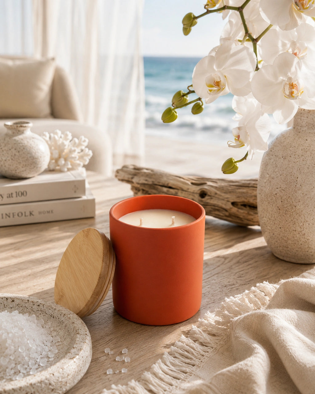 A terracotta orange candle jar with its wooden lid leaning beside it on a light wood table, styled in a bright coastal interior with white orchids, a vase, driftwood, and an ocean view in the background.