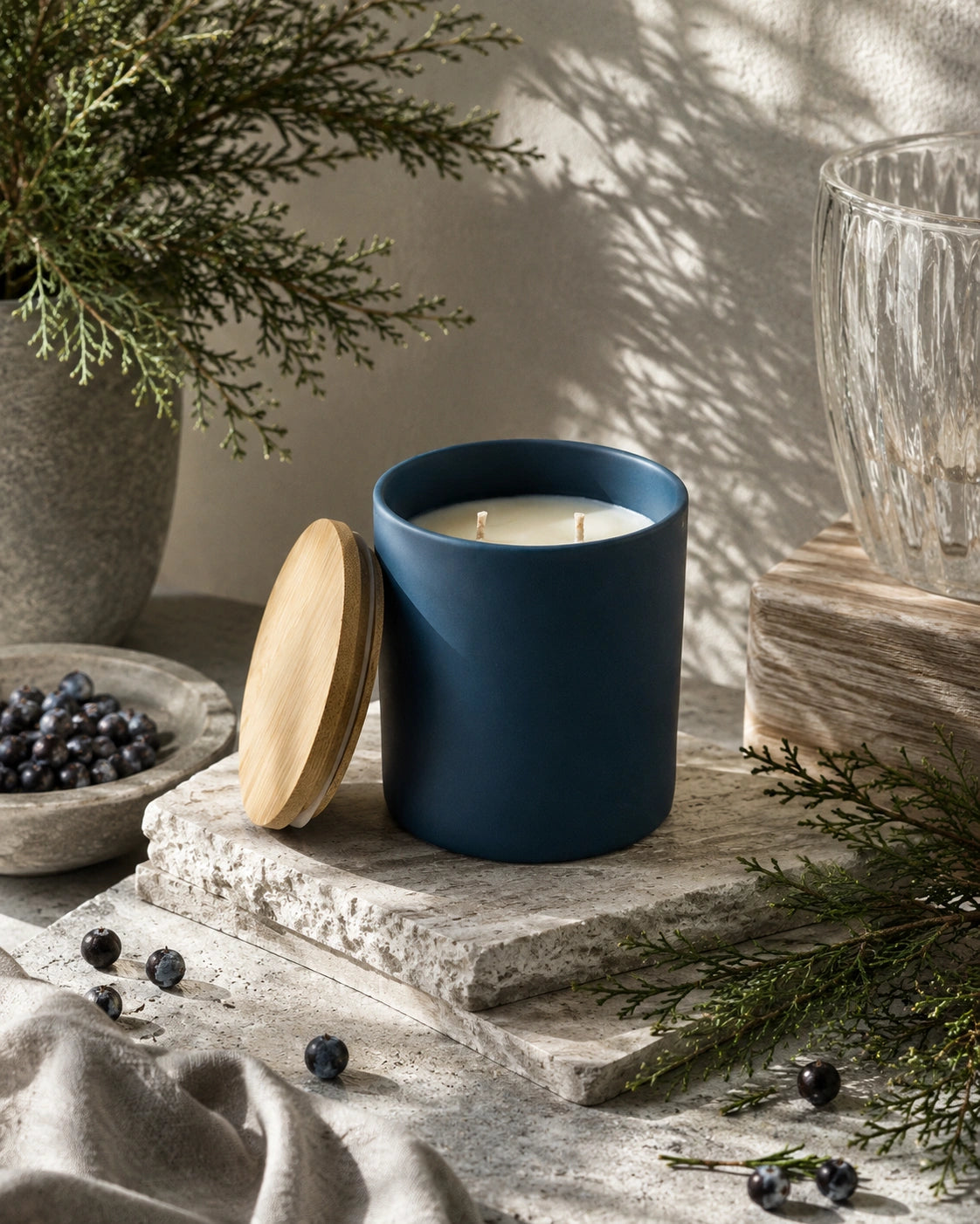 A matte navy blue candle jar with a light wood lid, styled on a rustic stone slab with pine branches, blueberries, and soft natural shadows.