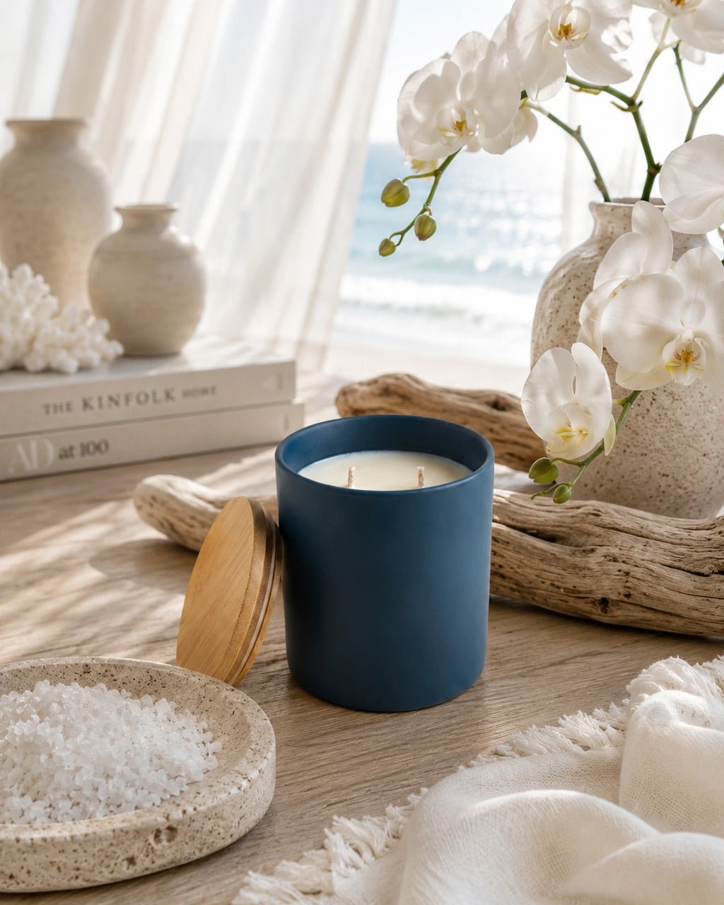 A matte navy blue candle jar with a light wood lid, styled on a wooden table with white bath salts, a textured vase of white orchids, and a soft white throw blanket in a bright, coastal-themed setting.