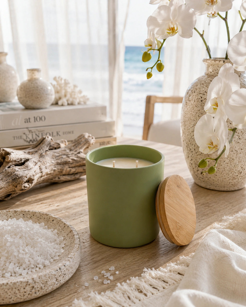 A sage green candle in a matte ceramic jar with its wooden lid resting beside it on a wooden table. The scene is styled with a speckled bowl of sea salt, driftwood, a vase of white orchids, and a window looking out to the ocean in the background.