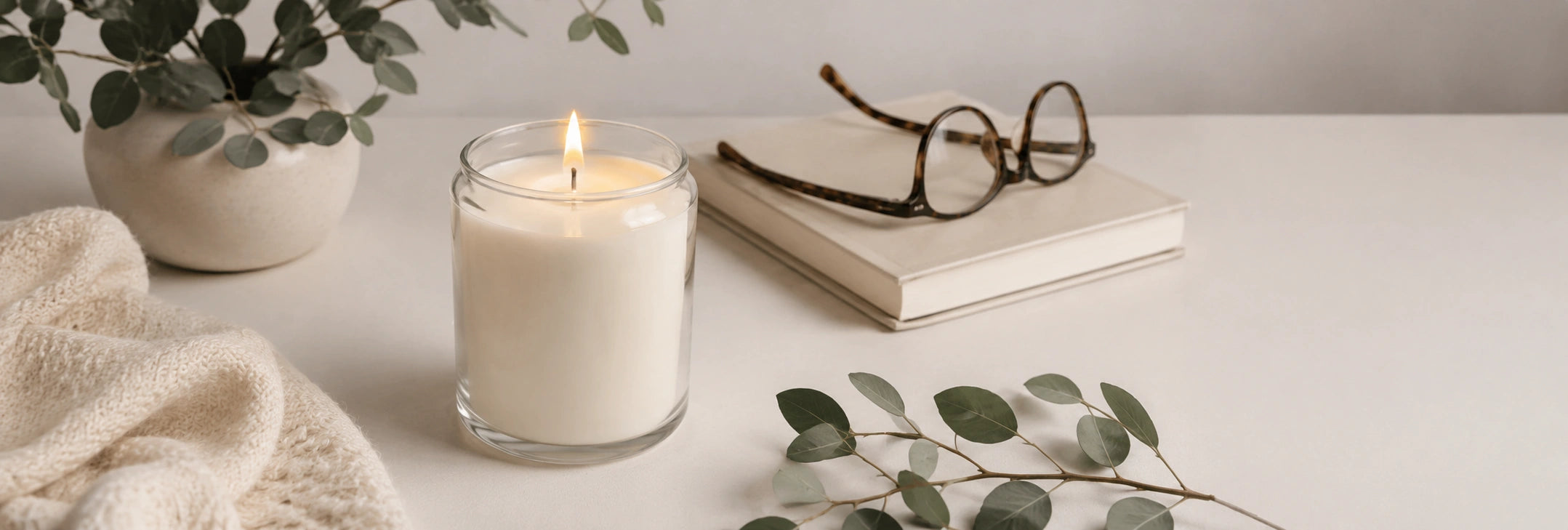 Candle in a glass holder with a book and glasses on a light surface, surrounded by greenery.