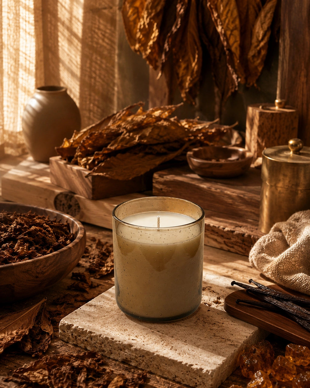 A lit candle in a clear glass jar sits on a stone slab in a rustic, warm-toned setting with dried leaves, wood, and natural textures.