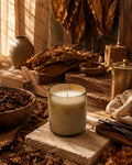 A lit candle in a clear glass jar sits on a stone slab in a rustic, warm-toned setting with dried leaves, wood, and natural textures.