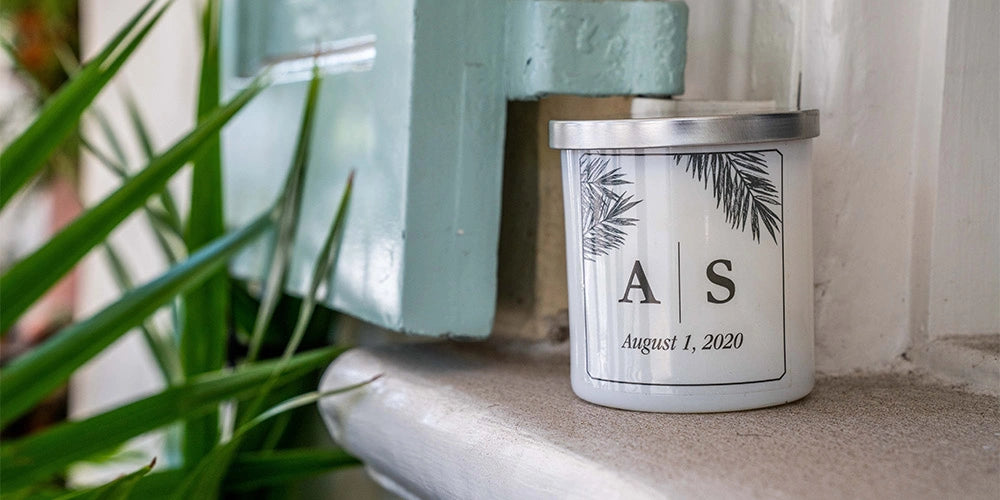 Candle with 'AS' initials and date on a surface with a plant in the background