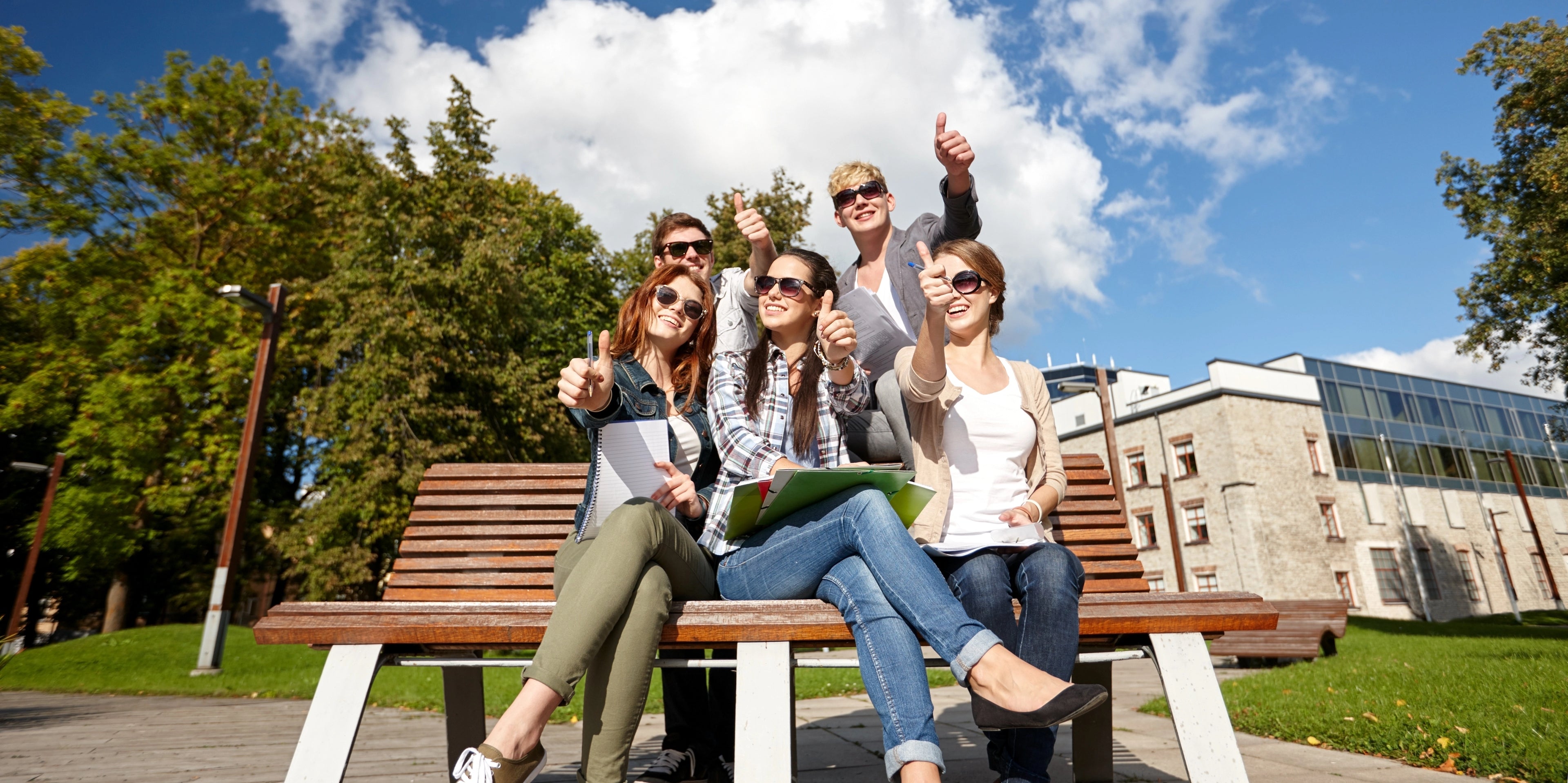 Group of students sitting on a bench outdoors with a building and trees in the background