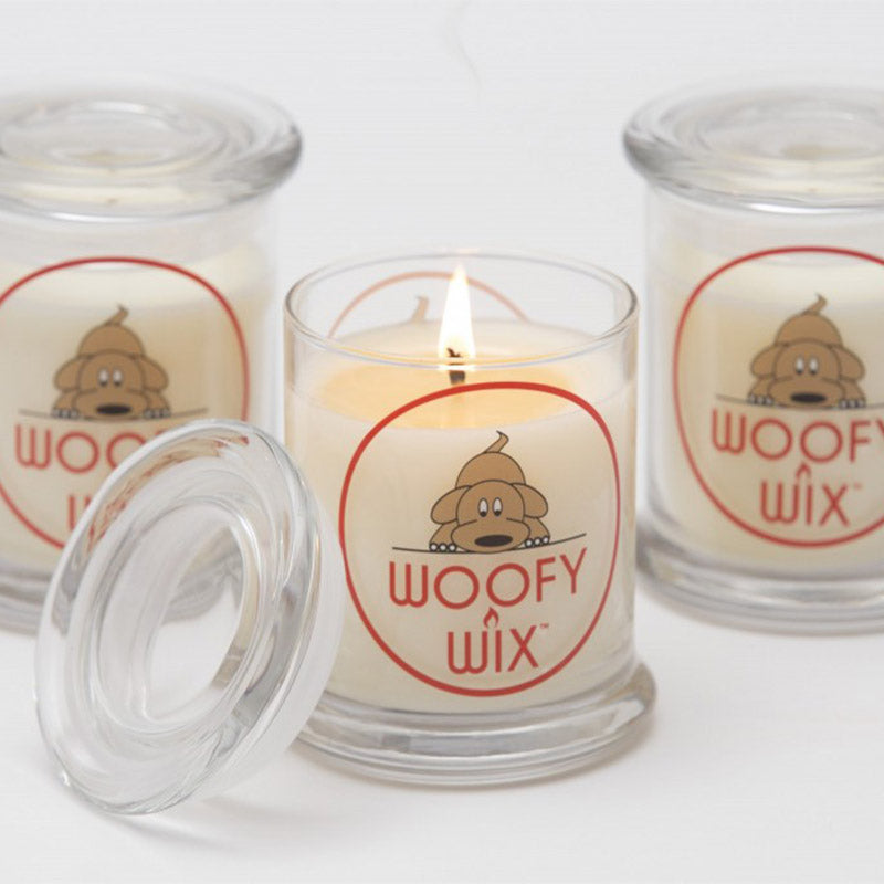Three clear glass candles with a label reading 'WOOFY WIX', featuring a cartoon dog illustration, and a lit candle in the foreground.